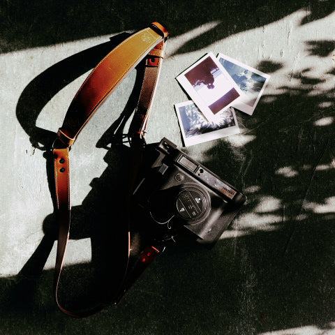 Krützberg Zeithalter MONO Kameragurt aus Wasserbüffel- und Rindsleder, flach arrangiert neben einer analogen Kamera und Polaroid-Fotos im natürlichen Licht.